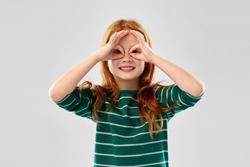 childhood and people concept - smiling red haired girl in green striped shirt having fun and looking through finger glasses over grey background