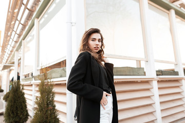 Pretty young elegant woman in a T-shirt in a trendy black coat in jeans is standing near a modern summer cafe and enjoys a spring day. Beautiful girl for a walk. New collection of women's clothing.