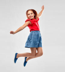 childhood, fun and motion concept - happy smiling girl in red shirt and skirt jumping over grey background