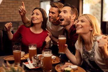 Young cheerful people having fun while watching sports match on TV in pub.
