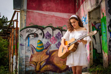 Young woman dressed in white holds a classical guitar in front of an abandoned building full of graffiti