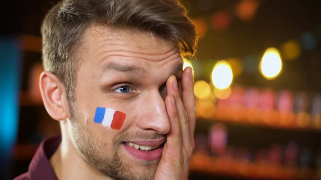 Upset bearded football fan with french flag on cheek making facepalm team losing