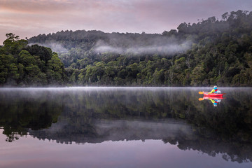 A girl enjoys a tranquil paddle in a kayak at dawn down a flat river, with misty rainforest and...