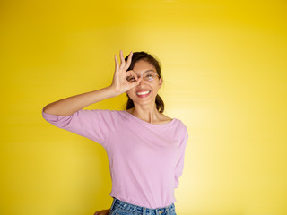 Beautiful young Asian woman show OK sign over her eye isolated on yellow background