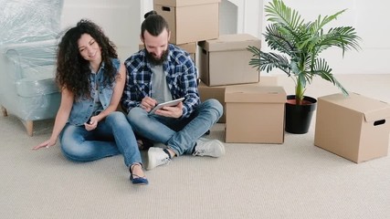 Young couple having rest after moving stuff to their new apartment, sitting on floor, sending housewarming invitation.