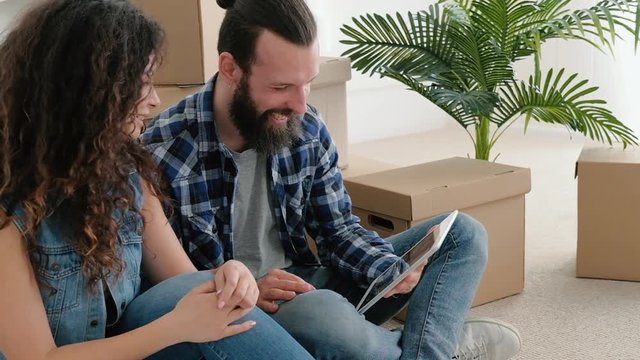 Young couple having fun after moving stuff to their new apartment, sitting on floor, watching comedy show, laughing.