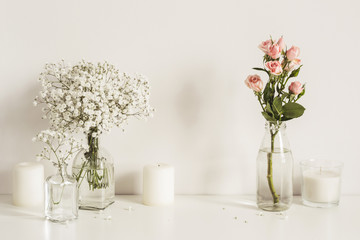 Composition with white and pink flowers in glass bottles and candles on table wall background. Copy space for artwork