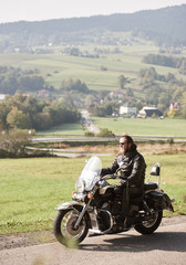 Fototapeta na wymiar Long-haired bearded cool rider in sunglasses and black leather clothing riding cruiser powerful motorcycle along sunny asphalt road on bright summer day on background of green rural misty landscape.