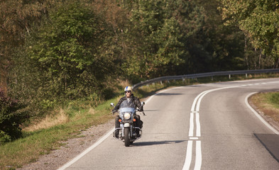 Bearded motorcyclist in helmet, sunglasses and black leather clothing riding bike along empty asphalt road with white marking on bright sunny summer day on background of green dense woody forest.