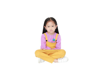 Portrait of angry little Asian child girl in pink-yellow pink-yellow dungarees sitting isolated on white background with copy space.