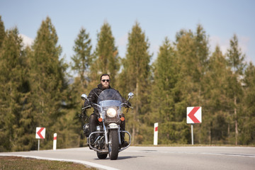 Alone motorcyclist in black leather outfit and sunglasses riding cruiser motorbike along beautiful road on bright sunny summer day