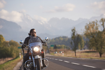 Obraz na płótnie Canvas Portrait of handsome bearded biker in black leather jacket holding motorcycle handles on country roadside on blurred background of green landscape, distant white mountain peaks and bright sky.