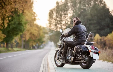 Foto auf Acrylglas Motorrad Rückansicht eines hübschen bärtigen Motorradfahrers in schwarzer Lederjacke und Sonnenbrille, der auf einem Cruiser-Motorrad sitzt, auf verschwommenem Hintergrund einer geraden Straße, die sich bis zum Horizont und goldenen Herbstbäumen erstreckt.