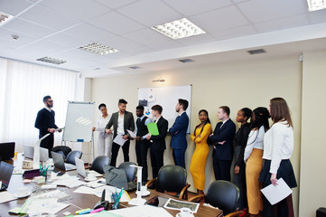 Mixed race business coach presenting report standing near whiteboard pointing on sales statistic shown on diagram and chart teach diverse company members gathered together in conference room.