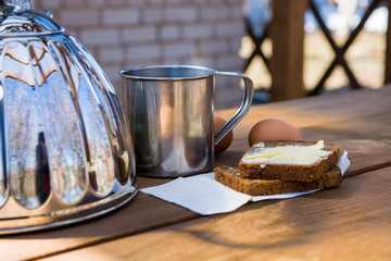On the wooden table is a kettle and a stainless steel mug, next to a sandwich with butter and eggs. Poured in a mug of strong tea. Concept: lunch in nature.