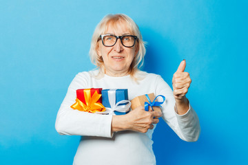 Old woman with a happy facial expression is holding gifts and showing thumbs up gesture against a blue background. Mothers day concept, congratulations, birthday or holiday, approval and gesture Ok.