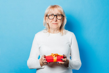 Old woman holding a gift in her hands against a blue background. Mother's Day, congratulation, birthday and holiday concept.