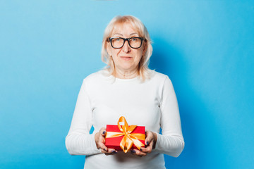 Old woman holding a gift in her hands against a blue background. Mother's Day, congratulation, birthday and holiday concept.
