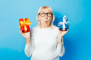 An old woman with a surprised and admired facial expression holds gifts in her hands against a blue background. Mother's Day, congratulation, birthday and holiday concept.