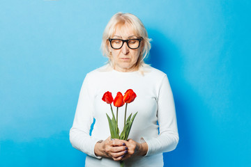 Old woman with a happy facial expression is holding a bouquet of flowers on a blue background. Mother's day concept, congratulation, birthday and holiday, the arrival of spring.