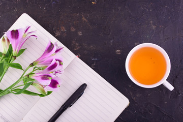 Fragrant tea in a white cup, beautiful flowers, a black background, a notebook with a pen and glasses.