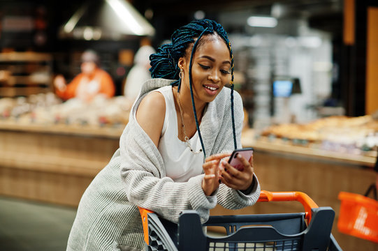 African american woman with shopping cart trolley in the supermarket store look on mobile phone.