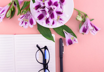 Fragrant tea in a white cup, beautiful flowers, a pink background, a notebook with a pen and glasses.