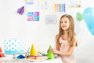 adorable smiling kid at festive table with party caps and birthday cake looking at camera