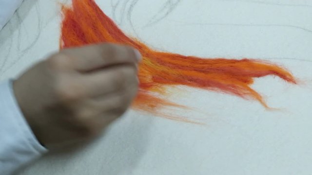 Close-up. A teenager in a white shirt pokes a needle over an orange dry felt. Making a picture of colored wool on a white fabric basis. Handicraft craft.