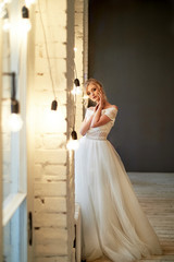 A beautiful petite blonde is standing by the window. The bride in a lace dress. Bride's morning