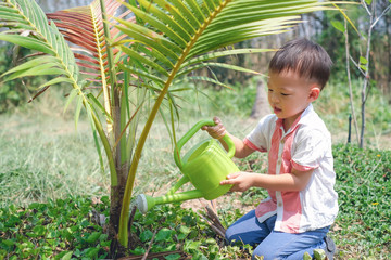 Cute smiling happy little Asian 2 - 3 years old toddler baby boy child watering young tree with watering can on black soil in the green garden, Save the world and environment concept
