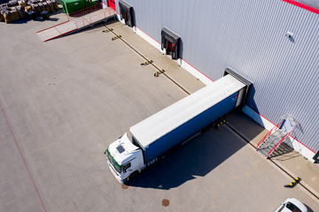 Aerial Shot of Truck with Attached Semi Trailer Leaving Industrial Warehouse/ Storage Building/ Loading Area where Many Trucks Are Load/ Unload Merchandise