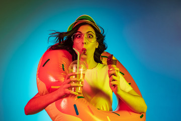 Obraz na płótnie Canvas Happy young woman in a rubber ring with an icecream and drink on trendy blue neon studio background. Female portrait. Concept of human emotions, facial expression, summer holidays or weekend.