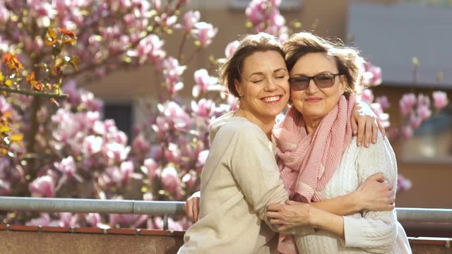 Mature mother and adult daughter hugging standing on a balcony on a background of flowering tree of magnolia. Bright spring sun, Mother's Day