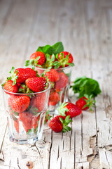 Organic red strawberries in two glasses and mint leaves on rustic wooden background.