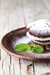 Fresh chocolate dark muffin with sugar powder and mint leaf on brown plate.