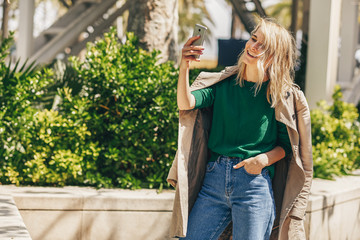 Young beautiful woman smiling brightly while holding smart phone strolling in the city at sunny day. Happy female making selfie. Pretty girl hold in hand cellphone, during conversation online