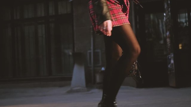Female leaving courtyard walking down street. Stylish urban woman. Red tartan skirt, legs in nylons patent boots closeup.
