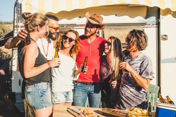 Group of cheerful people youthful young men and women have fun together in friendship with beers - old vintage alternative vacation caravan in background - rural place and guys laughing