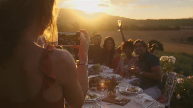 Group of young friends sitting around table and posing while a woman taking their picture with her mobile phone. Female taking photographs of friends at dinner party out in the yard.