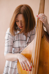 Beautiful young woman musician sitting on a vintage double bass on a beige background in a studio