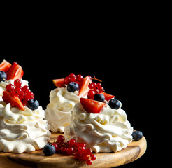Four homemade white mini desserts pavlova on wooden board with whipped cream, cut strawberries, blueberries, red currants 