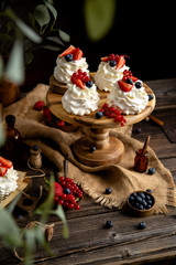 Four homemade white mini desserts pavlova on wooden cake stand with whipped cream and assorted berries on rustic table