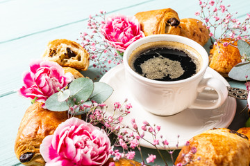 Cup of coffee, mini fresh croissants chocolate bun and carnation flowers on turquoise background.