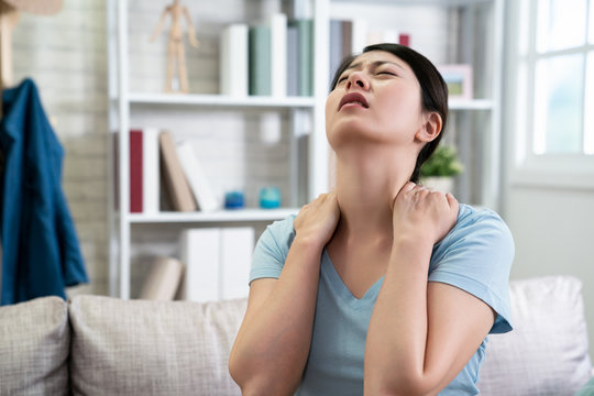 unhappy asian chinese housewife woman has neck pain stretching painful body frowning face. illness japanese wife at home sitting on couch with hurting shoulder. upset female feeling sick indoors.