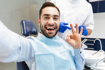 Image of brunette man sitting in dental chair at medical center and take selfie photo