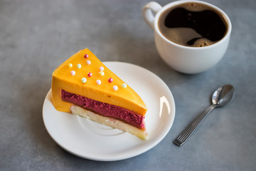 A piece of mousse cake with a taste of mango and strawberry on a white plate and a cup of coffee.