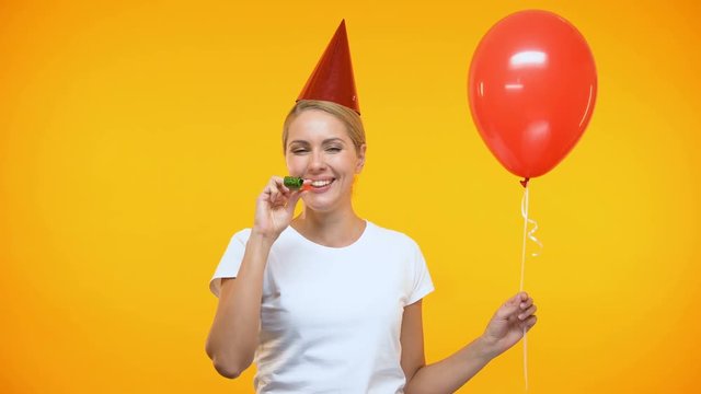 Cheerful young lady in party hat blowing horn, holding red balloon, birthday