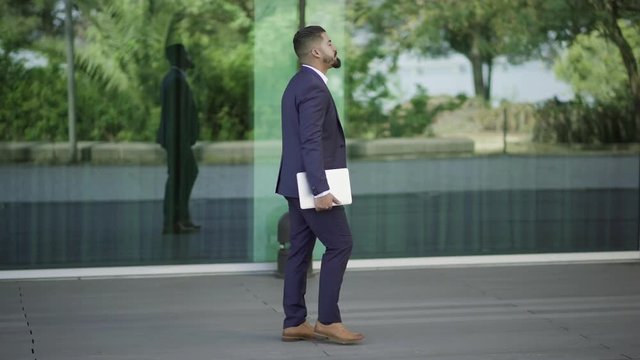 Businessman with laptop walking on street. Side view of bearded businessman holding laptop computer and going nar modern office building, tracking shot. Business concept
