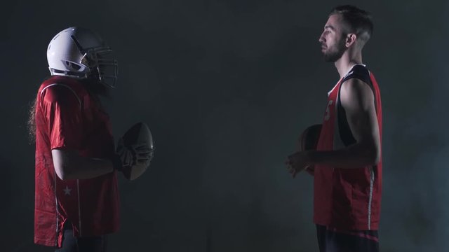 Two male sports players of different games standing in front of each other on the dark background. The american football player and basketball player look at each other holding balls. Side view